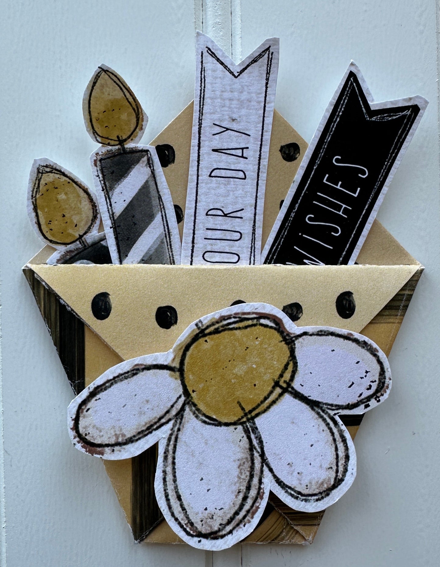 A handmade scrapbook paper embellishment featuring a white daisy with a gold center, black and white banners reading 'WISHES' and 'OUR DAY', and gold foil leaf accents on a white paneled background.