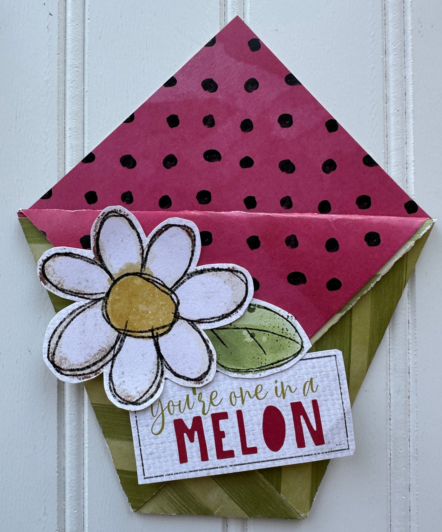 A handmade paper gift envelope or card holder with a pink polka-dot interior, a green patterned exterior, a white flower embellishment, and a small card that reads 'you're one in MELON' on a white paneled background.