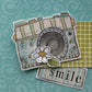 A decorative paper craft, handmade card or scrapbook design featuring a stylized vintage camera with floral details, a white daisy flower, and a small tag reading 'smile' on a patterned teal background.