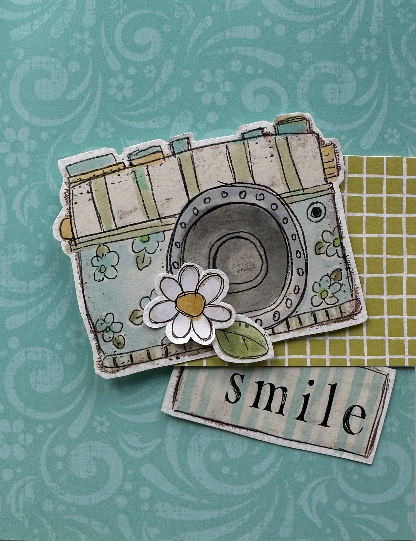 A decorative paper craft, handmade card or scrapbook design featuring a stylized vintage camera with floral details, a white daisy flower, and a small tag reading 'smile' on a patterned teal background.