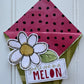 A handmade paper gift envelope or card holder with a pink polka-dot interior, a green patterned exterior, a white flower embellishment, and a small card that reads 'you're one in MELON' on a white paneled background.